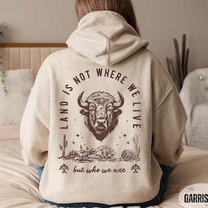May include: A beige hoodie with a graphic on the back featuring a bison head, desert plants, and the text "LAND IS NOT WHERE WE LIVE but who we are." The hoodie has a drawstring hood and long sleeves.
