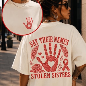 May include: An off-white t-shirt featuring a red graphic on the back with the text "SAY THEIR NAMES" and "STOLEN SISTERS" surrounding a handprint with a heart cutout, a rose, feathers, and a ribbon. A smaller matching handprint is on the front.