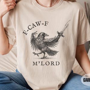 May include: Beige t-shirt featuring a detailed black and white illustration of a crow holding a sword. The text "F-CAW-F M'LORD" is printed above and below the crow. The Garrison Trend logo is visible.