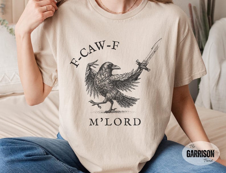 May include: Beige t-shirt featuring a detailed black and white illustration of a crow holding a sword. The text "F-CAW-F M'LORD" is printed above and below the crow. The Garrison Trend logo is visible.