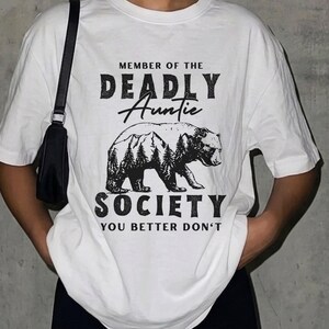 Può includere: T-shirt bianca con grafica nera. Il design presenta il testo "MEMBER OF THE DEADLY Auntie SOCIETY YOU BETTER DON'T" e un orso con una scena di montagna e foresta.