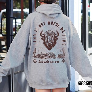 May include: A light gray hoodie with a brown graphic of a bison head, desert landscape, and the text "LAND IS NOT WHERE WE LIVE but who we are". The hoodie has a drawstring hood and long sleeves.