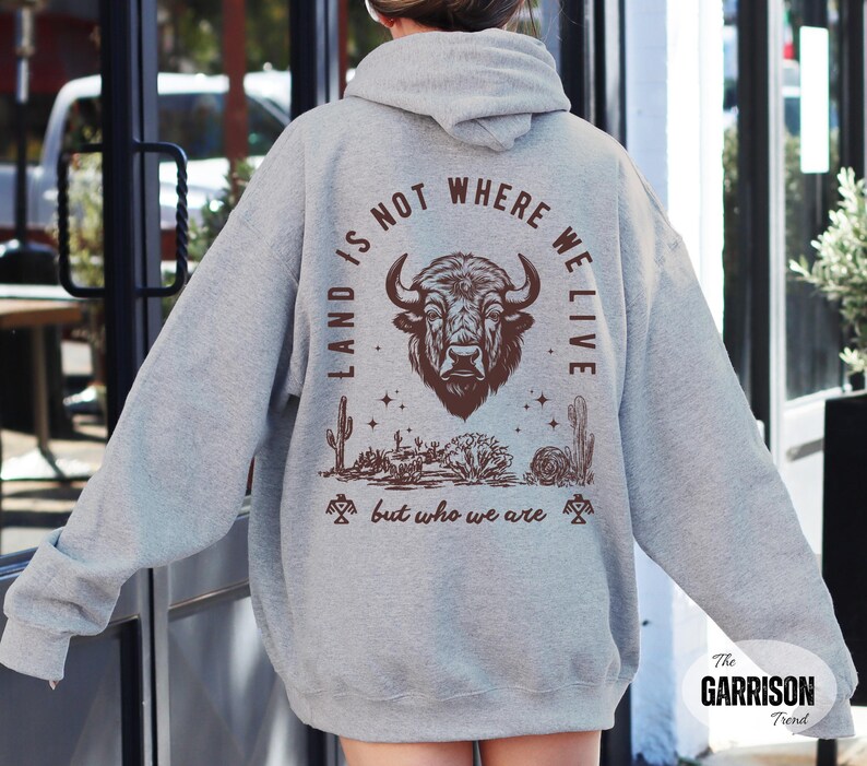 May include: A light gray hoodie with a brown graphic of a bison head, desert landscape, and the text "LAND IS NOT WHERE WE LIVE but who we are". The hoodie has a drawstring hood and long sleeves.