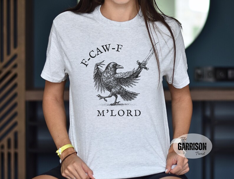 May include: Light gray t-shirt featuring a detailed black illustration of a crow holding a sword, with the text "F-CAW-F M'LORD" above and below the bird. The shirt has a crew neck and short sleeves.