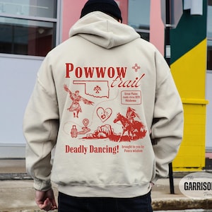 Puede incluir: Sudadera con capucha beige con un diseño gráfico rojo. El diseño incluye el texto "Powwow trail", ilustraciones de un bailarín nativo americano, un mapa de Oklahoma y un jinete a caballo. También se incluye el texto "Deadly Dancing!".