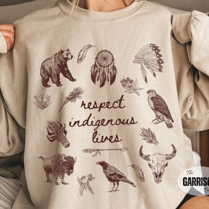 May include: Beige sweatshirt with brown illustrations of a bear, dreamcatcher, eagle, buffalo, and other Native American symbols. The words "respect indigenous lives" are printed on the front.