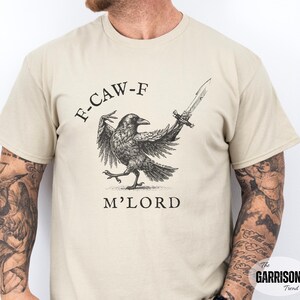 May include: Beige t-shirt featuring a black and white illustration of a raven holding a sword, with the text "F-CAW-F M'LORD" above and below the raven. The Garrison Trend logo is in the bottom right corner.