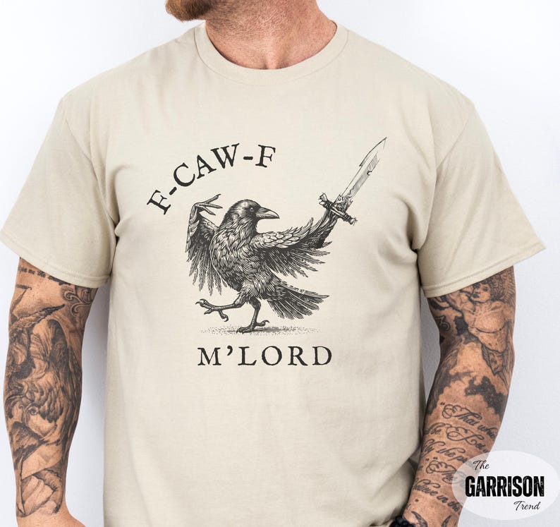 May include: Beige t-shirt featuring a black and white illustration of a raven holding a sword, with the text "F-CAW-F M'LORD" above and below the raven. The Garrison Trend logo is in the bottom right corner.