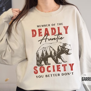 May include: Cream-colored sweatshirt with the text "MEMBER OF THE DEADLY Auntie SOCIETY YOU BETTER DON'T" in red and black. A brown bear graphic is centered below the text. The Garrison Trend logo is in the bottom right corner.