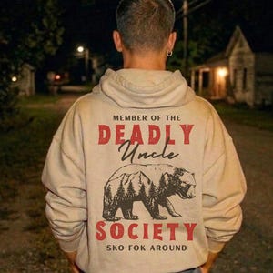 May include: Beige hoodie with the text "MEMBER OF THE DEADLY Uncle SOCIETY" and a bear graphic. The text and graphic are in red and black. The hoodie is worn by a person standing outside at night.