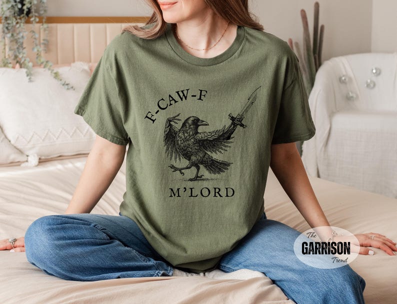 May include: Olive green t-shirt featuring a black crow illustration holding a sword, with the text "F-CAW-F M'LORD". The shirt is a comfortable fit and made of soft material.