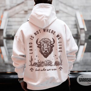 May include: White hoodie with a graphic on the back featuring a bison head, desert plants, and the text "LAND IS NOT WHERE WE LIVE but who we are."