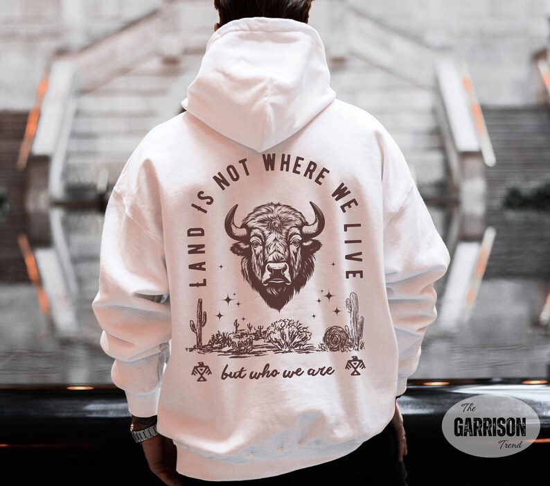 May include: White hoodie with a graphic on the back featuring a bison head, desert plants, and the text "LAND IS NOT WHERE WE LIVE but who we are."