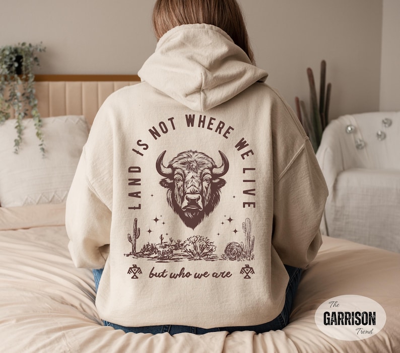 May include: A beige hoodie with a graphic on the back featuring a bison head, desert plants, and the text "LAND IS NOT WHERE WE LIVE but who we are." The hoodie has a drawstring hood and long sleeves.