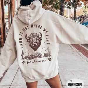 May include: Cream-colored hoodie with a graphic on the back featuring a bison head, cacti, and the text "LAND IS NOT WHERE WE LIVE BUT WHO WE ARE." The hoodie has a drawstring hood and long sleeves.