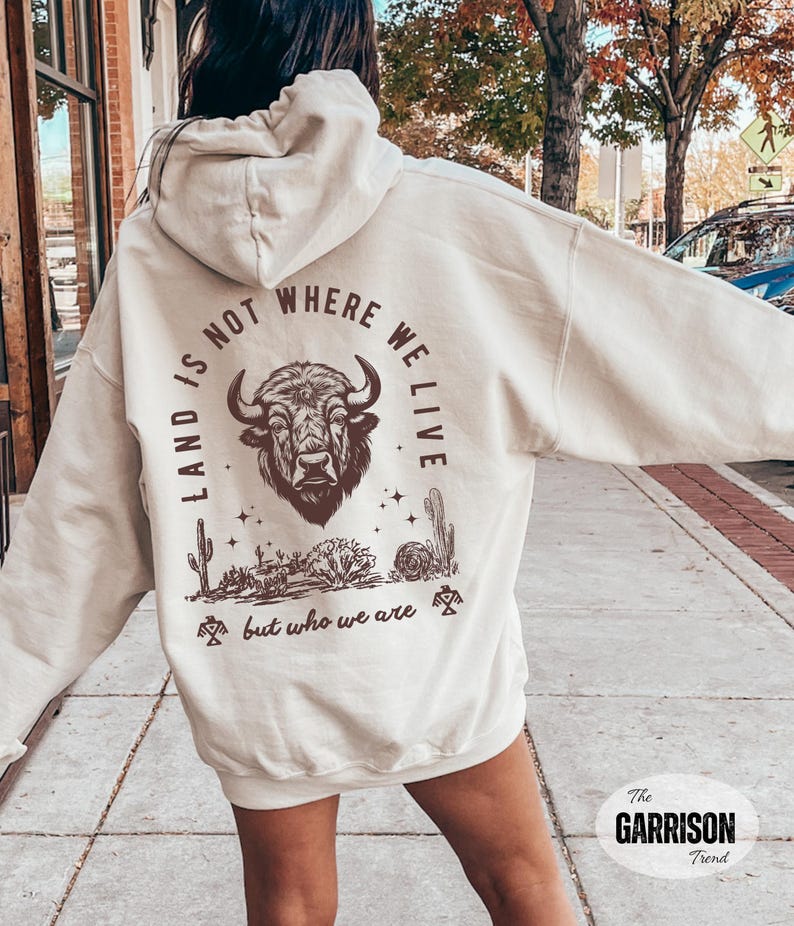 May include: Cream-colored hoodie with a graphic on the back featuring a bison head, cacti, and the text "LAND IS NOT WHERE WE LIVE BUT WHO WE ARE." The hoodie has a drawstring hood and long sleeves.