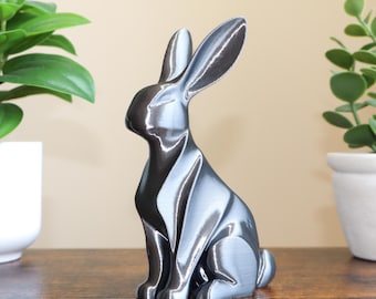 May include: A silver-colored rabbit figurine, posed in a seated position, with long ears and a smooth, reflective surface. The bunny statue is placed on a wooden surface, with green plants in white pots on either side.