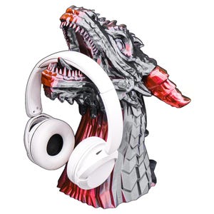 Puede incluir: Un soporte para auriculares con forma de dragón en tonos grises, rojos y negros, que sostiene un par de auriculares blancos. La cabeza del dragón está detallada con dientes afilados y escamas texturizadas. Los auriculares son de tipo circumaural.
