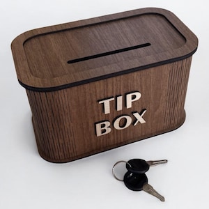Op de afbeelding: Een donkerbruine houten fooienbox met een gleuf en de tekst "TIP BOX". De box heeft een zwarte rand en wordt geleverd met twee sleutels.