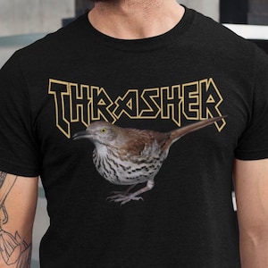 以下が含まれることがあります： 黒いTシャツ。金色の文字で「THRASHER」と書かれ、茶色の鳥の精巧なイラストが描かれています。鳥の胸には斑点があります。Tシャツは半袖で、背景はニュートラルです。