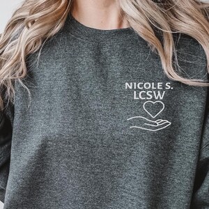 Könnte beinhalten: Dunkelgraues Sweatshirt mit dem Text "NICOLE S. LCSW" über einer weißen Grafik eines Herzens in einer Hand. Das Model trägt das Sweatshirt und zeigt das Peace-Zeichen mit den Fingern.