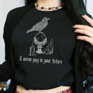 May include: Black long-sleeved shirt with a graphic of a raven perched on a crystal ball with candles on either side. The text "I sense gay in your future" is printed below the graphic.