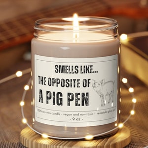 Puede incluir: Una vela de cera de soja marrón en un tarro de vidrio transparente con una etiqueta blanca que dice "Smells like... The opposite of a pig pen". La vela está encendida y tiene una pequeña ilustración de un cerdo en la etiqueta. La vela está sobre un posavasos de madera.