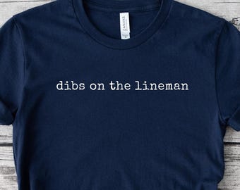 Op de afbeelding: Marineblauw t-shirt met de witte tekst "dibs on the lineman". Het shirt heeft opgerolde mouwen en is aan de onderkant geknoopt. Het woord "NAVY" wordt weergegeven in een marineblauwe cirkel. Het t-shirt is gemaakt van zacht materiaal.