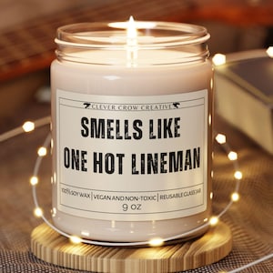 May include: A lit candle in a clear glass jar with a label that reads "SMELLS LIKE ONE HOT LINEMAN." The candle sits on a small wooden coaster. The text "CLEVER CROW CREATIVE" is above the main text. The candle is 9 oz.