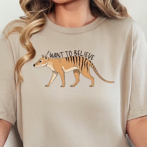 Op de afbeelding: Tan-kleurig T-shirt met een cartoon illustratie van een Tasmaanse tijger met zwarte strepen. De tekst "I WANT TO BELIEVE" staat boven het dier. Het shirt is een neutrale kleur en heeft korte mouwen.