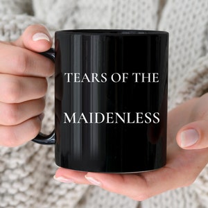 May include: A black ceramic mug with white text that reads "Tears of the Maidenless".