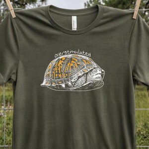 Könnte beinhalten: Ein militärgrünes Kurzarm-T-Shirt mit einer weißen und orangefarbenen Strichgrafik einer Schildkröte, die sich in ihrem Panzer versteckt. Das Wort "overstimulated" ist in einer weißen, handschriftlichen Schrift über der Grafik gedruckt.