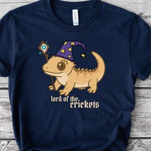May include: Navy blue t-shirt with a cartoon lizard in a wizard hat and staff, with the text "lord of the crickets". The lizard is beige and the hat is purple with yellow stars. The shirt is made of a soft material.