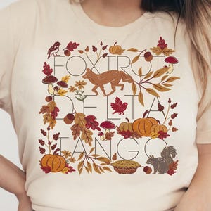 Può includere: T-shirt color crema con grafica a tema autunnale. Il design include una volpe, zucche, foglie, funghi e uno scoiattolo, con le parole "FOX TROT DELTA TANGO" in marrone, arancione e rosso.