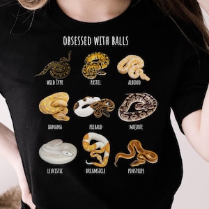 Puede incluir: Camiseta negra con la frase "OBSESSED WITH BALLS" e ilustraciones de varias mutaciones de serpientes, incluyendo Wild Type, Pastel, Albino, Banana, Piebald, Mojave, Leucistic, Dreamsicle y Pinstripe. La palabra "BLACK" está en un círculo en la parte inferior.