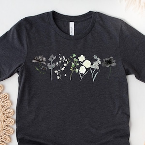 May include: Dark grey heather t-shirt with a floral design. The design features black, white, and green botanical illustrations. The shirt has a crew neck and short sleeves. The text "DARK GREY HEATHER" is in a circle at the bottom left.