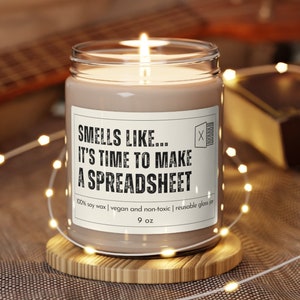 May include: A brown soy wax candle in a clear glass jar with a white label that reads "Smells Like... It's Time To Make A Spreadsheet". The candle is on a wooden coaster and surrounded by white string lights.