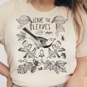 May include: A beige t-shirt with a black and white illustration of a bird, leaves, and acorns. The text "LEAVE THE LEAVES" is printed on the shirt.