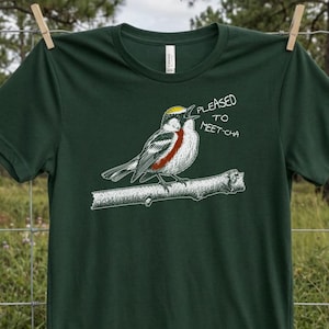 Puede incluir: Una camiseta de manga corta en color verde bosque oscuro con una ilustración gráfica de un pájaro posado en una rama. El pájaro tiene una corona amarilla y una mancha roja en el pecho. El texto "PLEASED TO MEET-CHA" aparece en blanco junto al pico.
