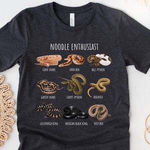Può includere: T-shirt grigio scuro melange con la scritta "NOODLE ENTHUSIAST" e illustrazioni di varie specie di serpenti, tra cui serpente del mais, boa della sabbia e pitone palla. La maglietta presenta anche serpente giarrettiera, pitone tappeto e serpente dal naso a porco.