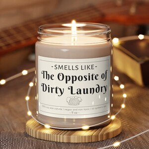 Puede incluir: Una vela de cera de soja marr&oacute;n en un tarro de vidrio transparente con una etiqueta blanca que dice "Smells Like - The Opposite of Dirty Laundry". La vela est&aacute; sobre un posavasos de madera y rodeada de luces blancas de hadas.