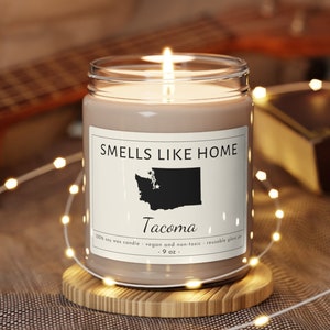 May include: A brown soy wax candle in a clear glass jar with a white label that reads "SMELLS LIKE HOME" and features a black silhouette of the state of Washington with the word "Tacoma" underneath. The candle is on a wooden coaster and surrounded by white string lights.