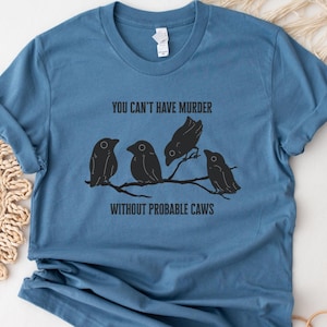 May include: Steel blue t-shirt with the text "YOU CAN'T HAVE MURDER WITHOUT PROBABLE CAWS" and a graphic of four black birds perched on a branch. The shirt has rolled-up sleeves.