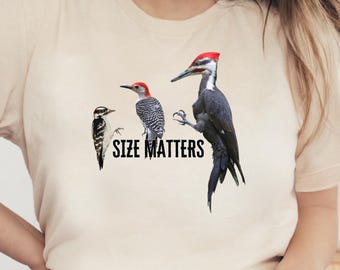 May include: A natural-colored t-shirt featuring three bird illustrations with the text "SIZE MATTERS" in black. The birds are a black and white woodpecker, a red-headed woodpecker, and a large black and white woodpecker with a red crest.