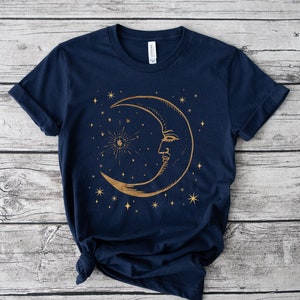 Pode incluir: Uma camiseta azul marinho com um design de lua crescente dourada e estrelas. A lua tem um rosto e as estrelas est&atilde;o espalhadas ao redor da lua.