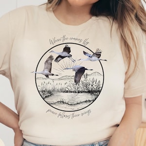 May include: A natural-colored t-shirt featuring a circular graphic with four cranes in flight over a landscape. The graphic includes the text "When the cranes fly, peace follows their wings."