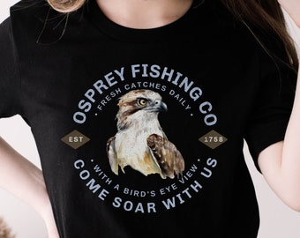 May include: Black t-shirt with a white circle graphic featuring an osprey with the text "Osprey Fishing Co. Fresh Catches Daily. Est 1758. With a Bird's Eye View. Come Soar With Us."