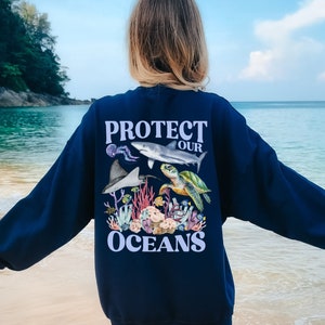 Könnte beinhalten: Ein marineblauer Sweatshirt mit einem grafischen Druck eines Hais, einer Meeresschildkröte und eines Korallenriffs. Der Text "PROTECT OUR OCEANS" ist in hellblau auf dem Sweatshirt gedruckt.