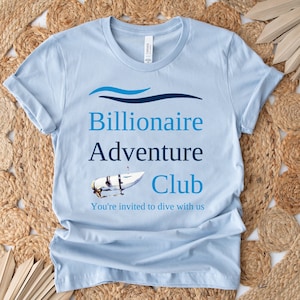 Może przedstawiać: Jasnoniebieska koszulka z napisem "Billionaire Adventure Club" i małym obrazkiem łodzi podwodnej. Tekst pod logo brzmi "You're invited to dive with us."
