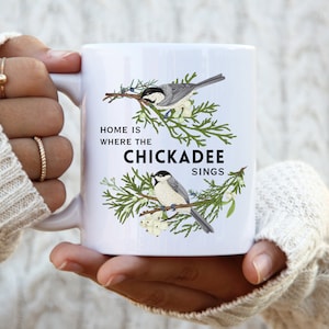 May include: White ceramic mug with a black and white illustration of two chickadees perched on a branch of a cedar tree. The text "Home is where the Chickadee sings" is printed on the mug.